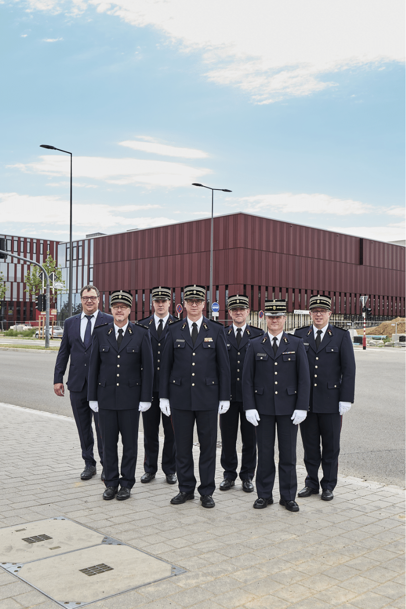 Groupe de sept hommes en uniforme, dont six portant des tenues de pompier et un en costume, posant ensemble devant un bâtiment moderne.