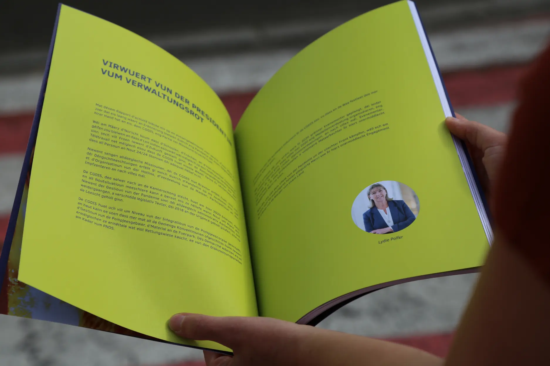 Une personne tenant ouverte une brochure avec des pages jaunes vives, contenant un texte en luxembourgeois et une photo ronde de Lydie Polfer.