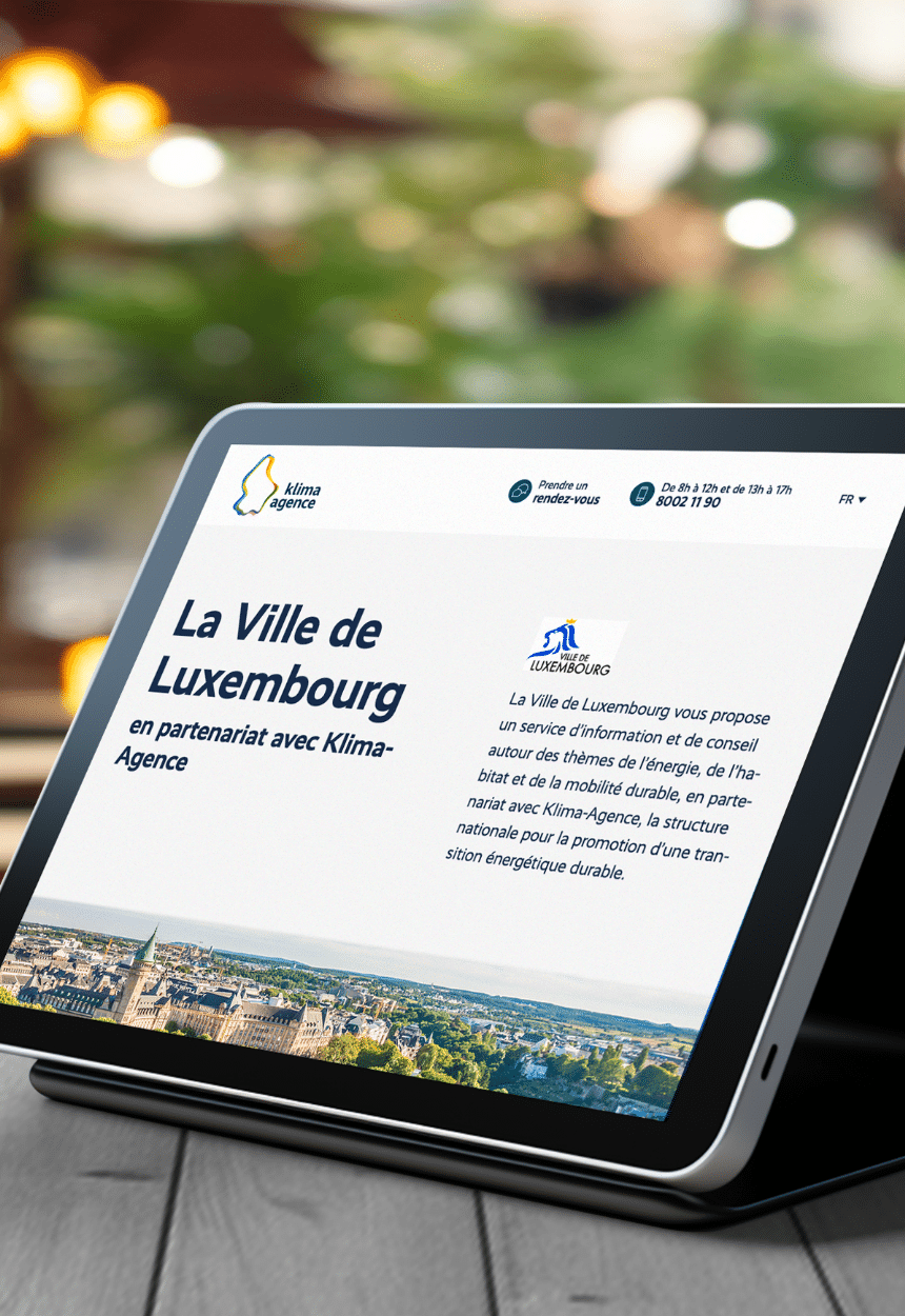 Tablette affichant une page web de la Ville de Luxembourg en partenariat avec Klima-Agence. Le texte indique un service d'information et de conseil sur les thèmes de l'énergie, de l'habitat et de la mobilité durable. En arrière-plan, une vue panoramique de la ville de Luxembourg est visible.