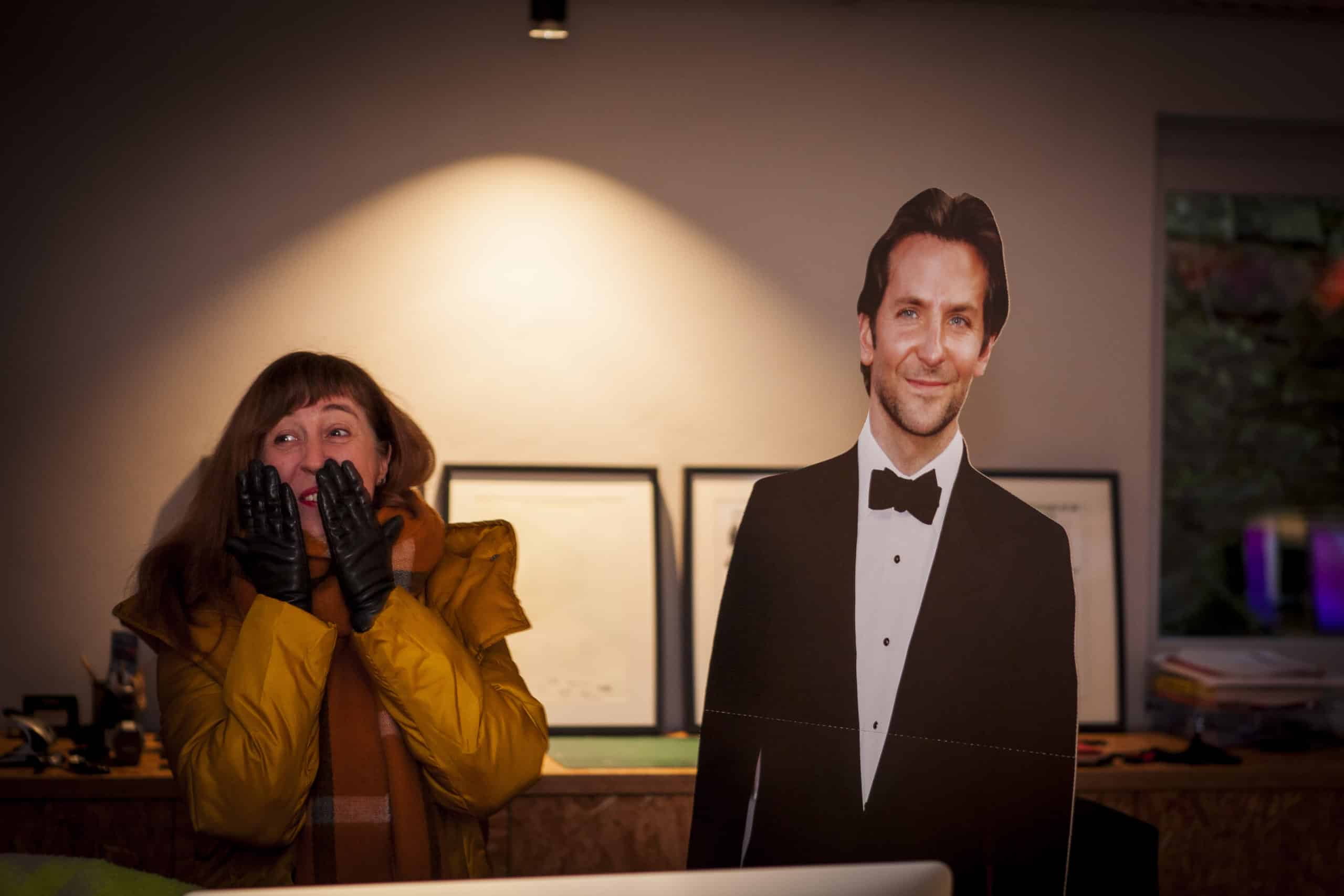 Une femme portant une veste jaune et une écharpe marron, debout à côté d'un carton découpé d'un homme en smoking noir et noeud papillon (Bradley Cooper), dans un bureau éclairé avec des cadres en arrière-plan. La femme a l'air surpris ou excité, couvrant son visage avec des gants noirs, ajoutant une touche amusante à la scène.
