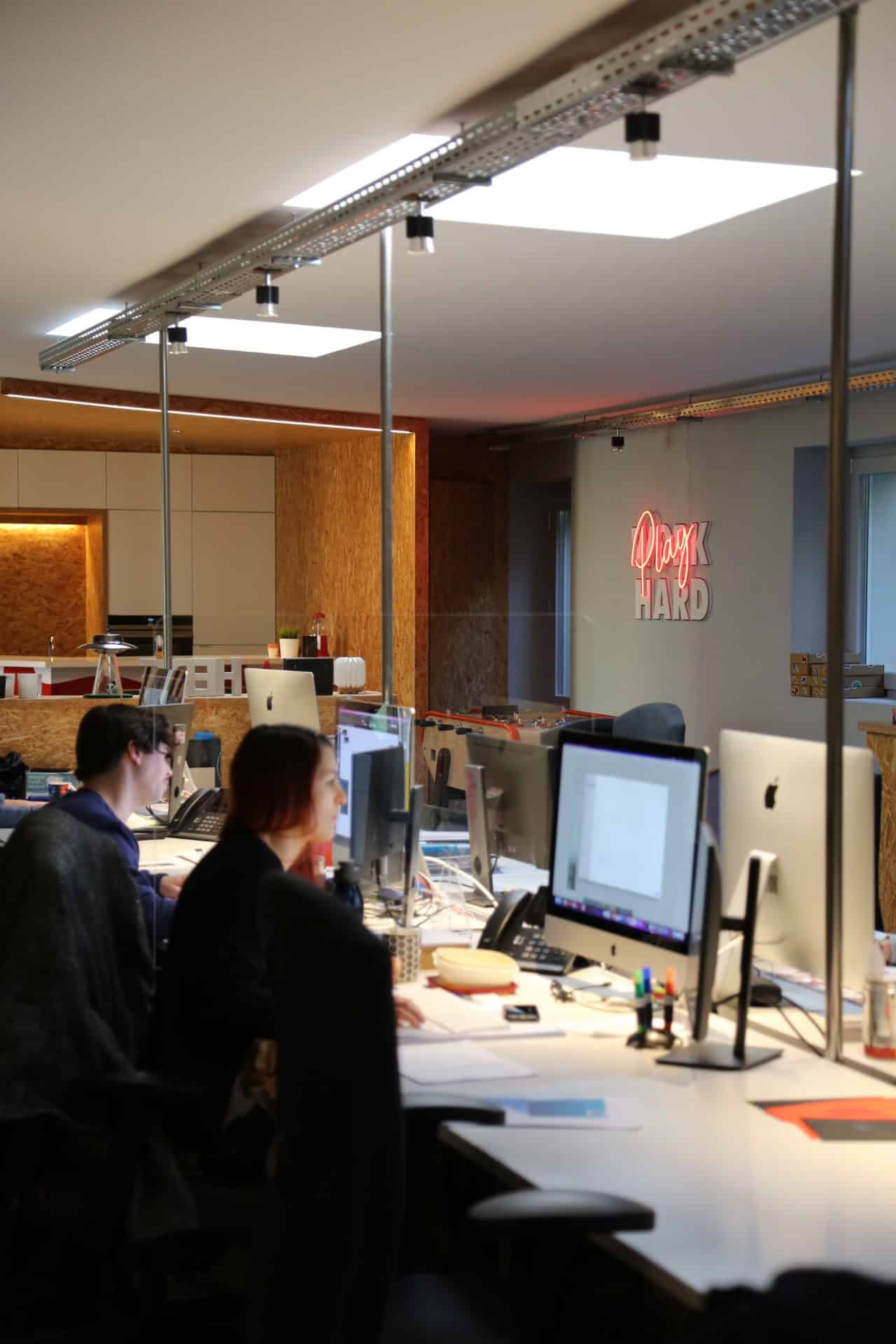Des employés de l'agence h2a travaillant dans un bureau moderne et lumineux. Plusieurs personnes sont assises devant des ordinateurs iMac, concentrées sur leur travail. Le bureau est divisé par des cloisons en verre, et on peut voir un panneau néon au fond indiquant 